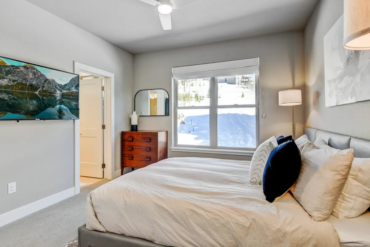 A serene bedroom scene featuring a comfortable bed, window views of the outdoors, a dresser, a large mirror, and a wall-mounted television. The tasteful decor and ample natural light create a peaceful retreat, ideal for unwinding with a movie or simply enjoying the mountain ambiance.