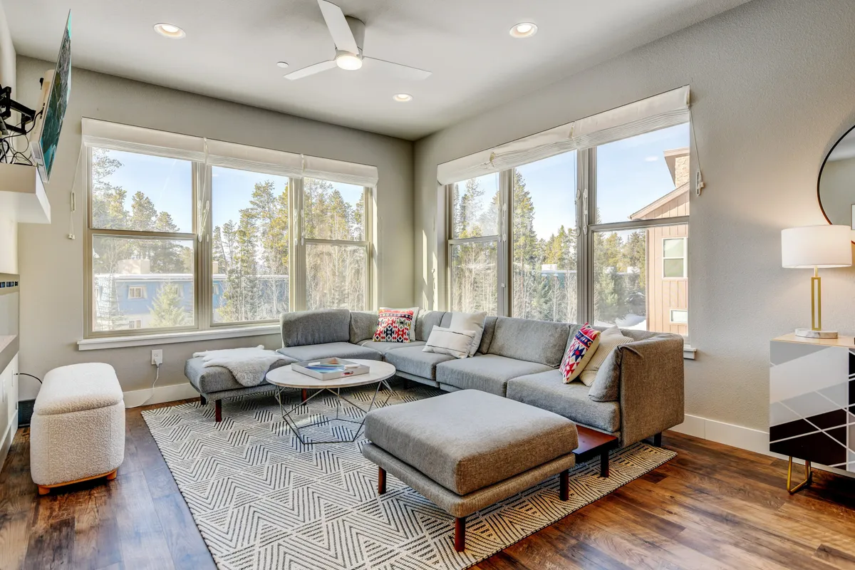 A spacious living area with a large sectional sofa, a low coffee table, and expansive windows offering tree views. Decorative pillows and a floor lamp add to the cozy, social vibe, making it an ideal spot for game nights or quiet relaxation.