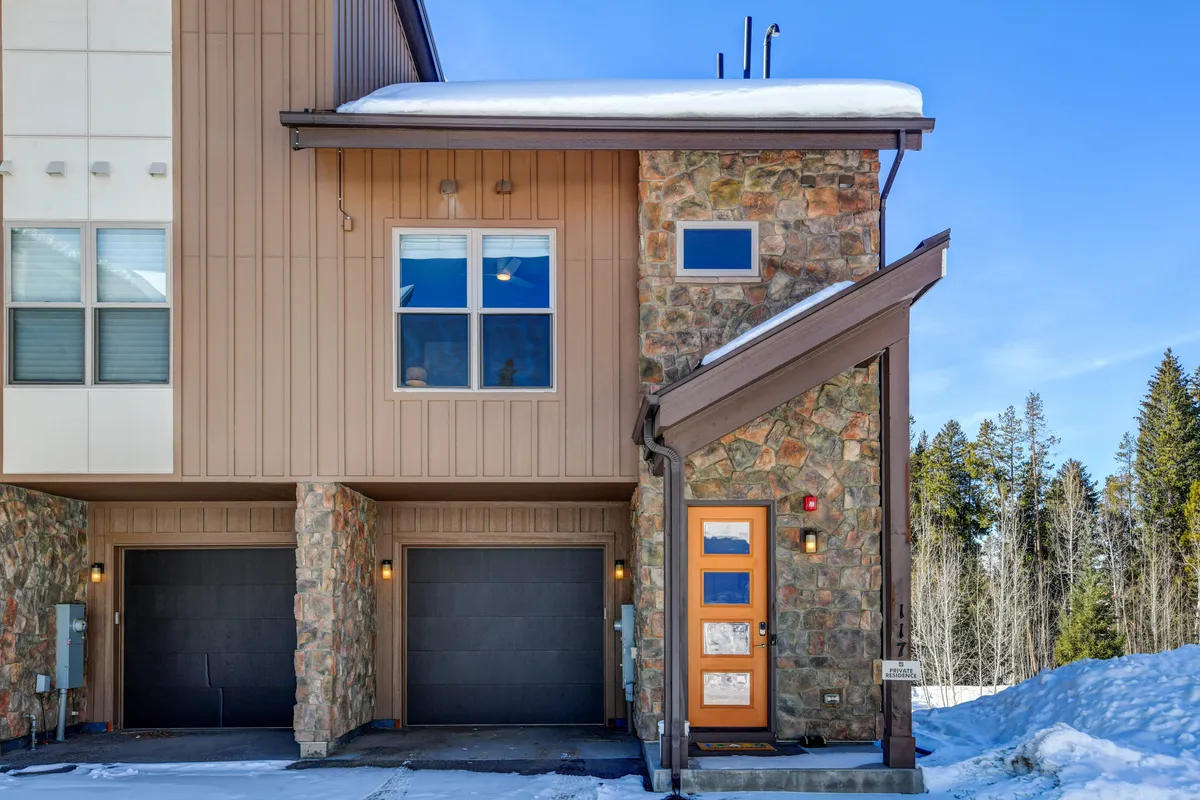 This exterior image shows a charming stone facade, a vibrant blue front door, and a snow-covered roof. The setting is welcoming and ideal for family (and pet) gatherings, with plenty of space out front for play or relaxation.