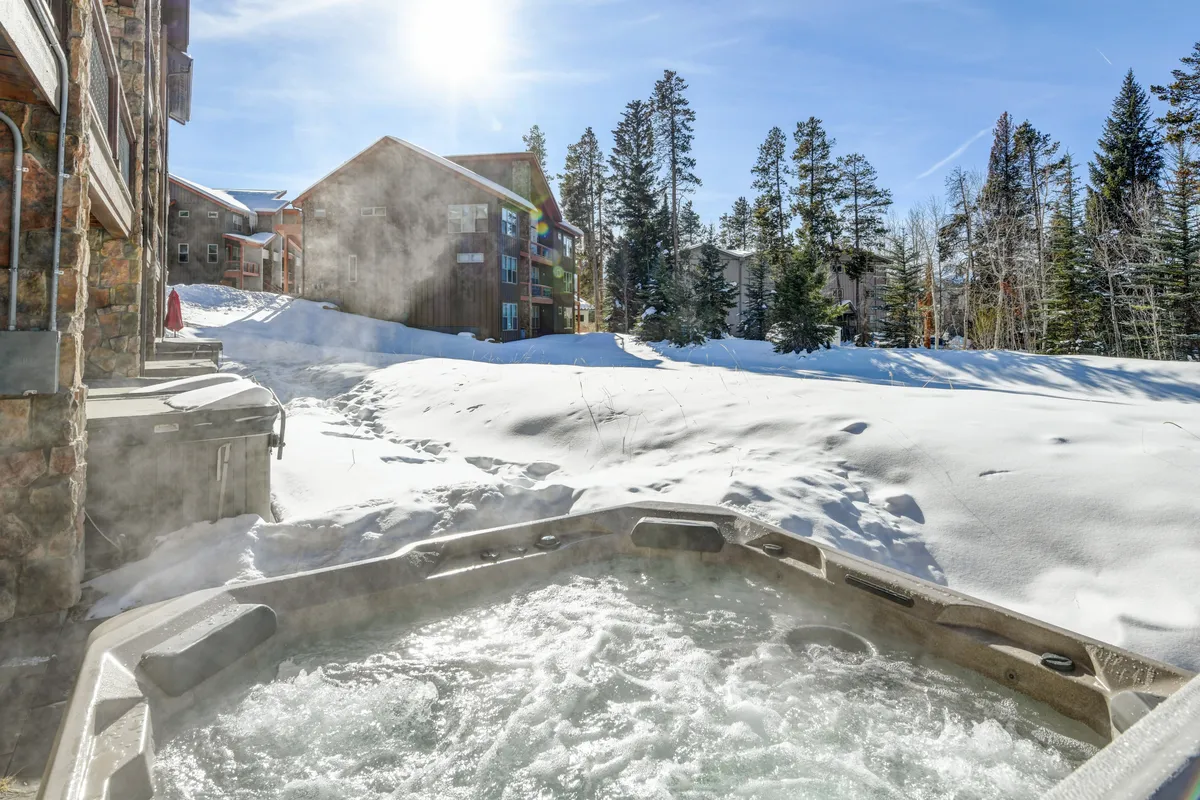 This exterior image features the inviting hot tub set against a backdrop of snow-covered landscape, tall trees, and buildings visible in the distance. The scene evokes both relaxation and adventure, with people enjoying a cozy soak while surrounded by natural beauty.