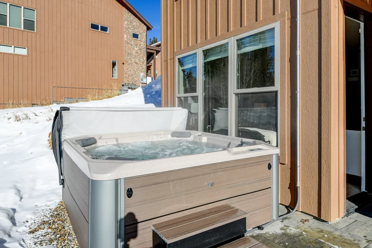 An exterior scene featuring a bubbling hot tub with steam rising, set beside large windows. The ground is blanketed in snow, and the view extends to the mountain landscape, emphasizing relaxation and outdoor leisure even in colder months.