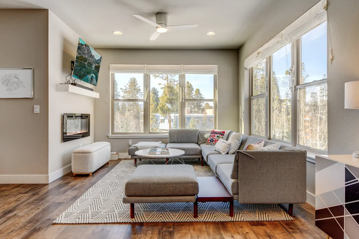 The image showcases a spacious, sunlit living room with a comfortable sectional sofa facing a modern fireplace and a large TV. Floor-to-ceiling windows offer breathtaking views of the Colorado landscape, while a stylish coffee table invites guests to gather for games or drinks. The mood is cozy and inviting, perfect for relaxing or entertaining with friends and family.