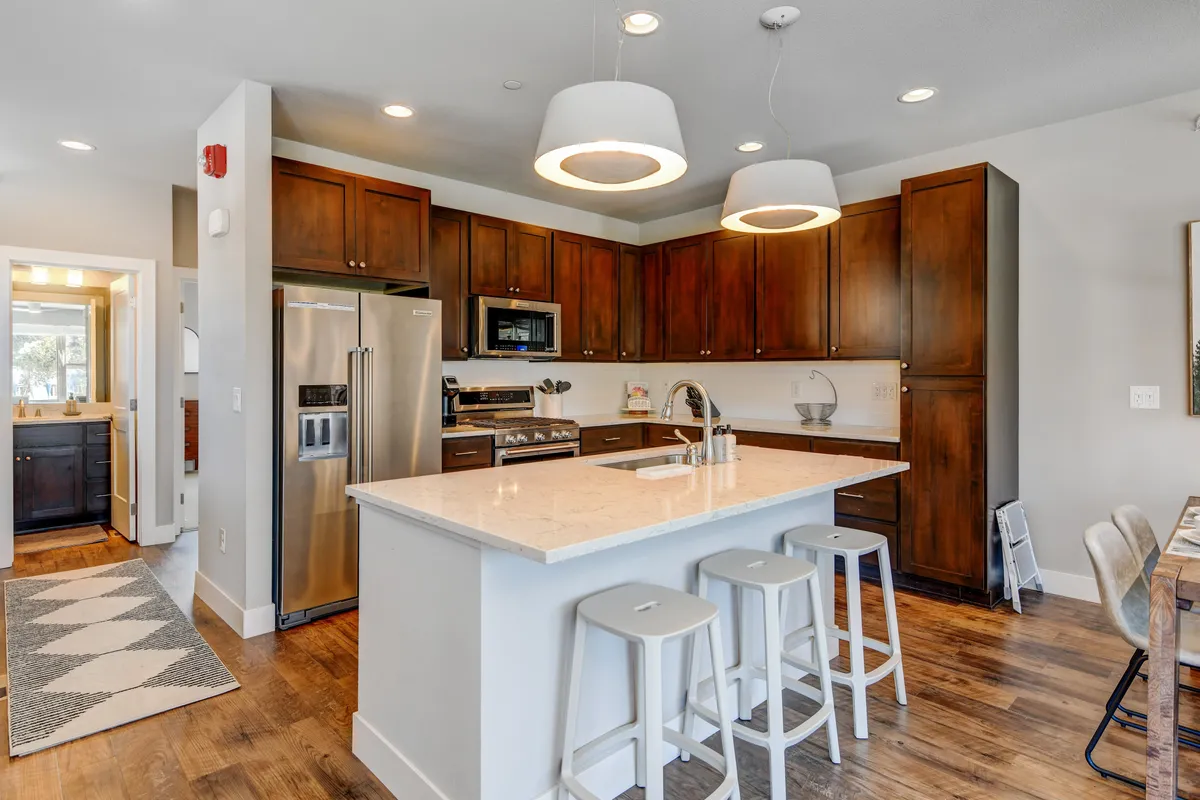 A warm, welcoming kitchen featuring a prominent kitchen island, sleek stainless steel appliances, rich wood cabinetry, and modern light fixtures. The open layout allows a direct view into the connected living and dining spaces, with subtle touches like premium cookware and a coffee station visible, setting a lively yet refined atmosphere.