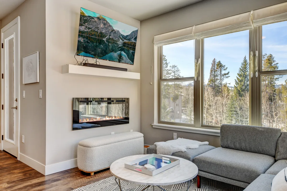 A cozy living room scene featuring a plush sofa, a coffee table, a wall-mounted television, and a unique gas fireplace. Natural light streams in through the windows, creating a warm and welcoming environment ideal for relaxing and socializing.