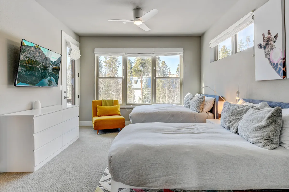 This image features two beds adorned with soft bedding, a bright yellow accent chair, large windows revealing views of nature, and a television mounted on the wall. The room feels airy and inviting, with touches that suggest both comfort and style. The arrangement is ideal for families or groups sharing a space, and the presence of the TV and natural views enhance the sense of retreat.