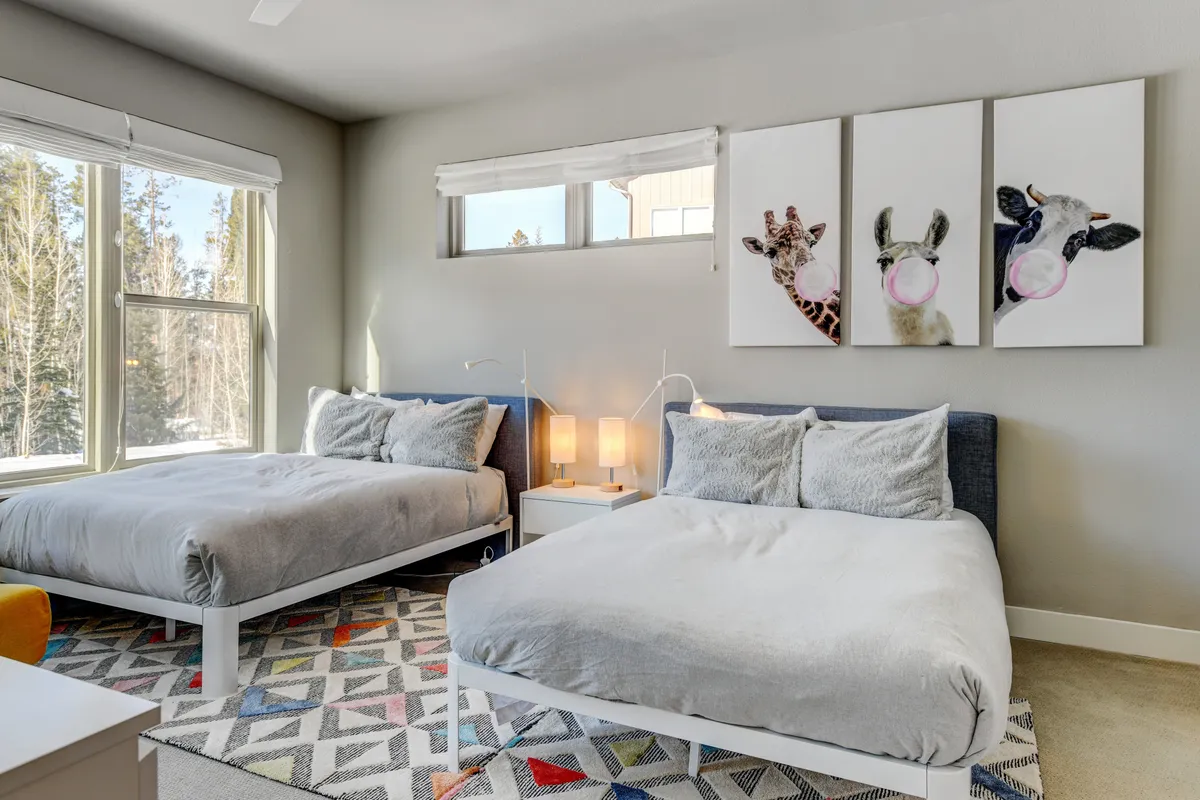 A cheerful bedroom with two beds, playful animal-themed artwork above the headboards, a colorful rug, and soft lighting from table lamps. Large windows flood the room with natural light, creating a bright and inviting space ideal for children or group accommodations.