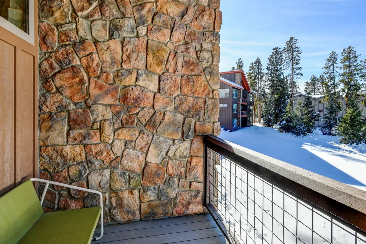 This exterior image showcases a stone wall, a green chair, and a snow-covered landscape dotted with tall pine trees. The tranquil outdoor space provides the perfect setting for relaxing, reading, or sipping coffee while surrounded by natural beauty.