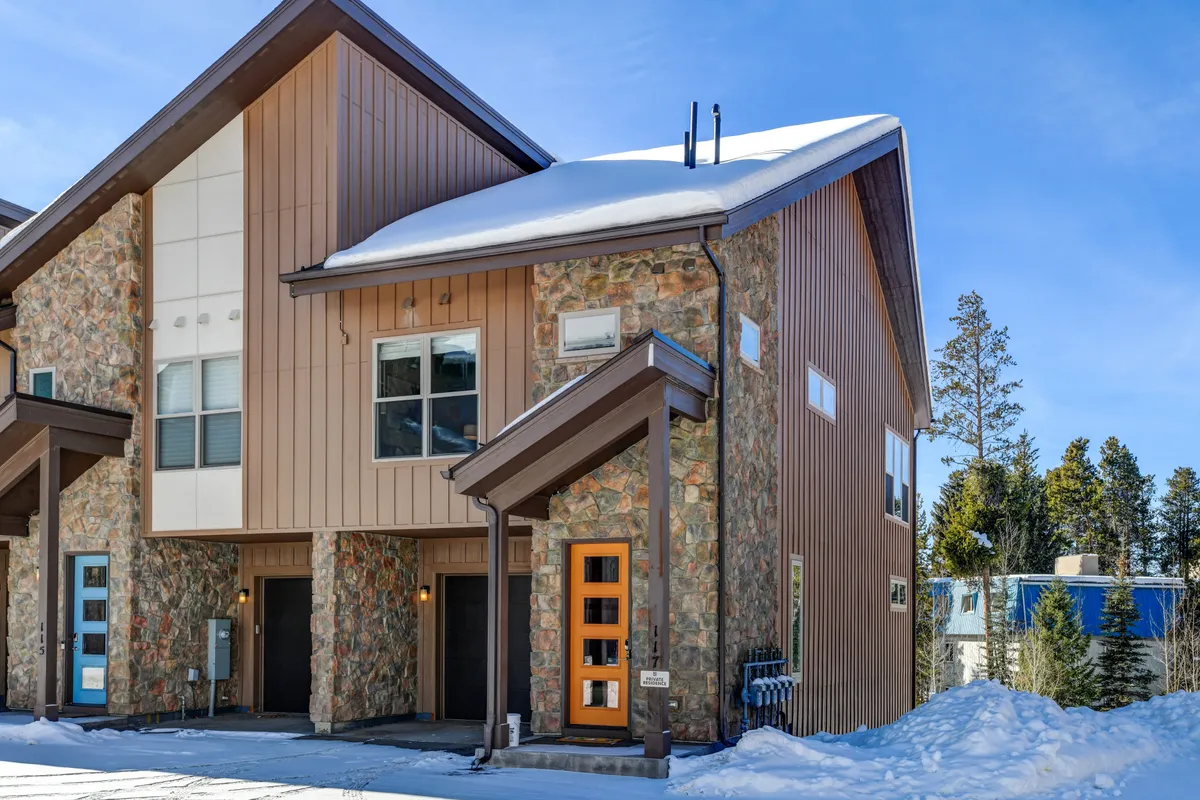 A welcoming exterior shot of the property, featuring a stone facade, colorful front doors, and a snowy landscape. The scene is vibrant with hints of activity, suggesting easy access to winter sports and outdoor gatherings right outside the home.
